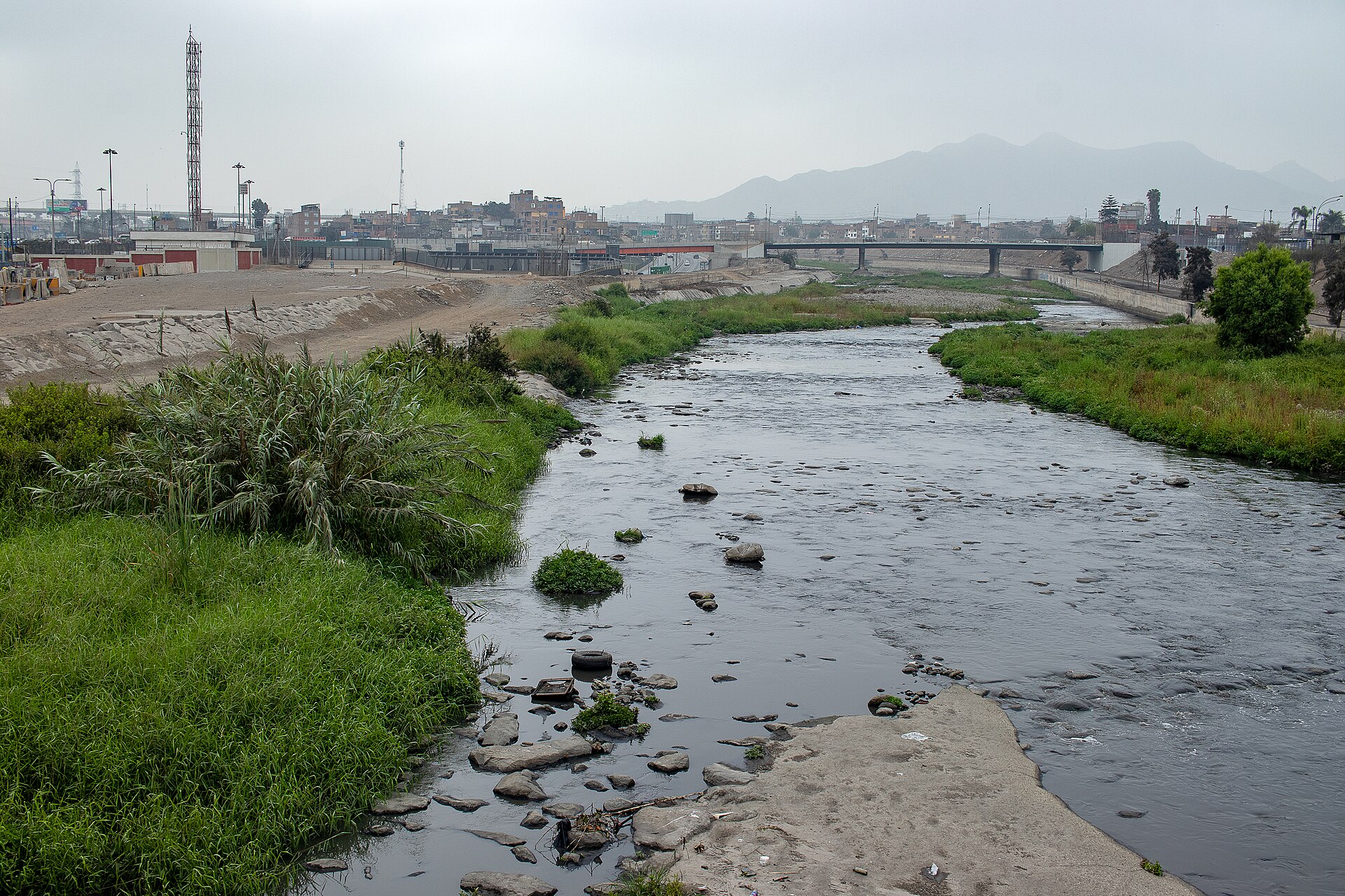 A shallow river with green banks flowing between low-rise buildings under a grey sky, with hills in the distance