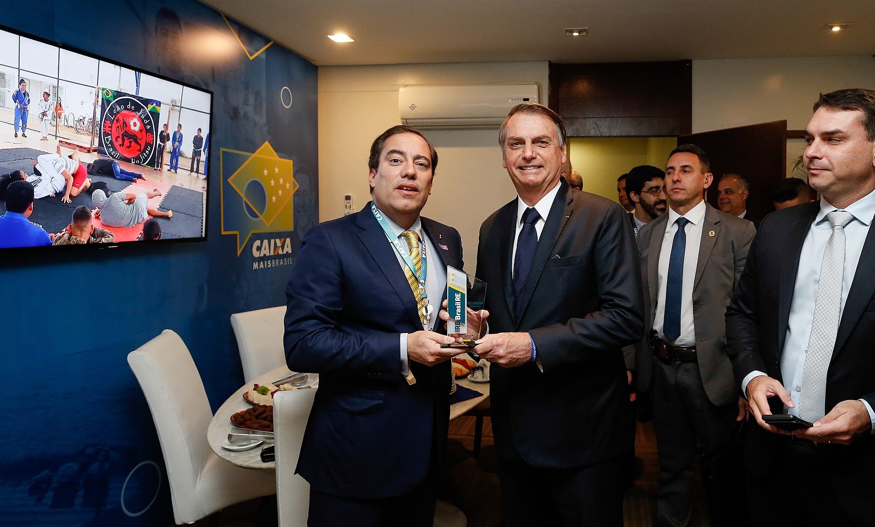 Jair Bolsonaro in a dark suit holds an award next to a man with a lanyard, while Flávio Bolsonaro stands on the right holding a phone