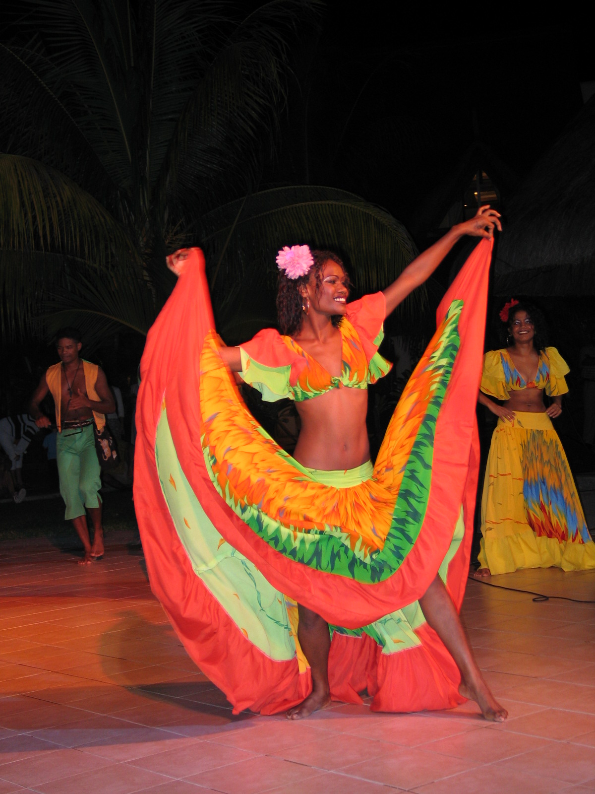 A sega dancer in a wide orange, green and yellow skirt performs at night under palm trees in Mauritius.