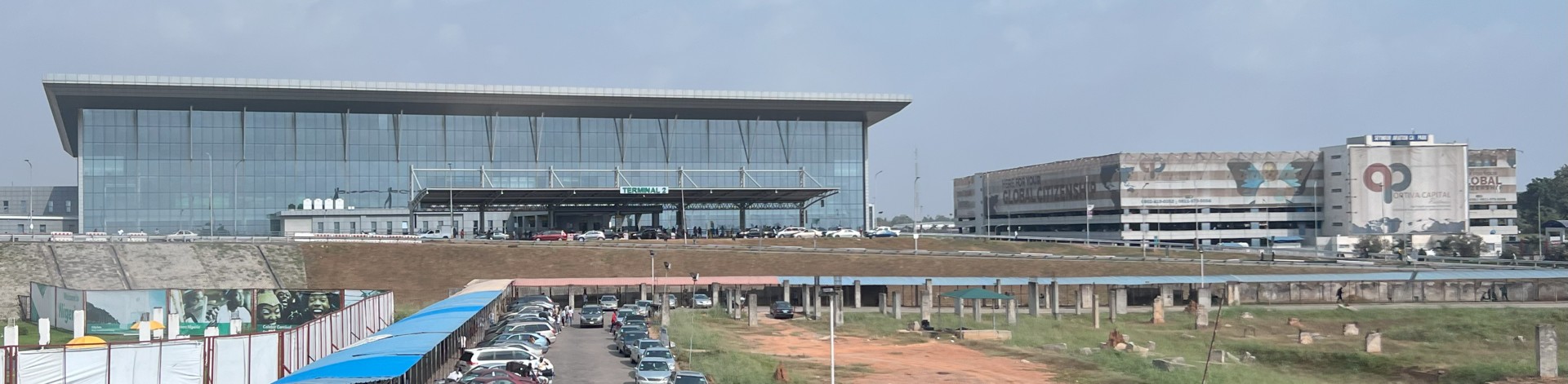 Glass-fronted Terminal 2 building at Lagos airport beside a multi-storey car park, with a parking lot and site hoardings in the foreground