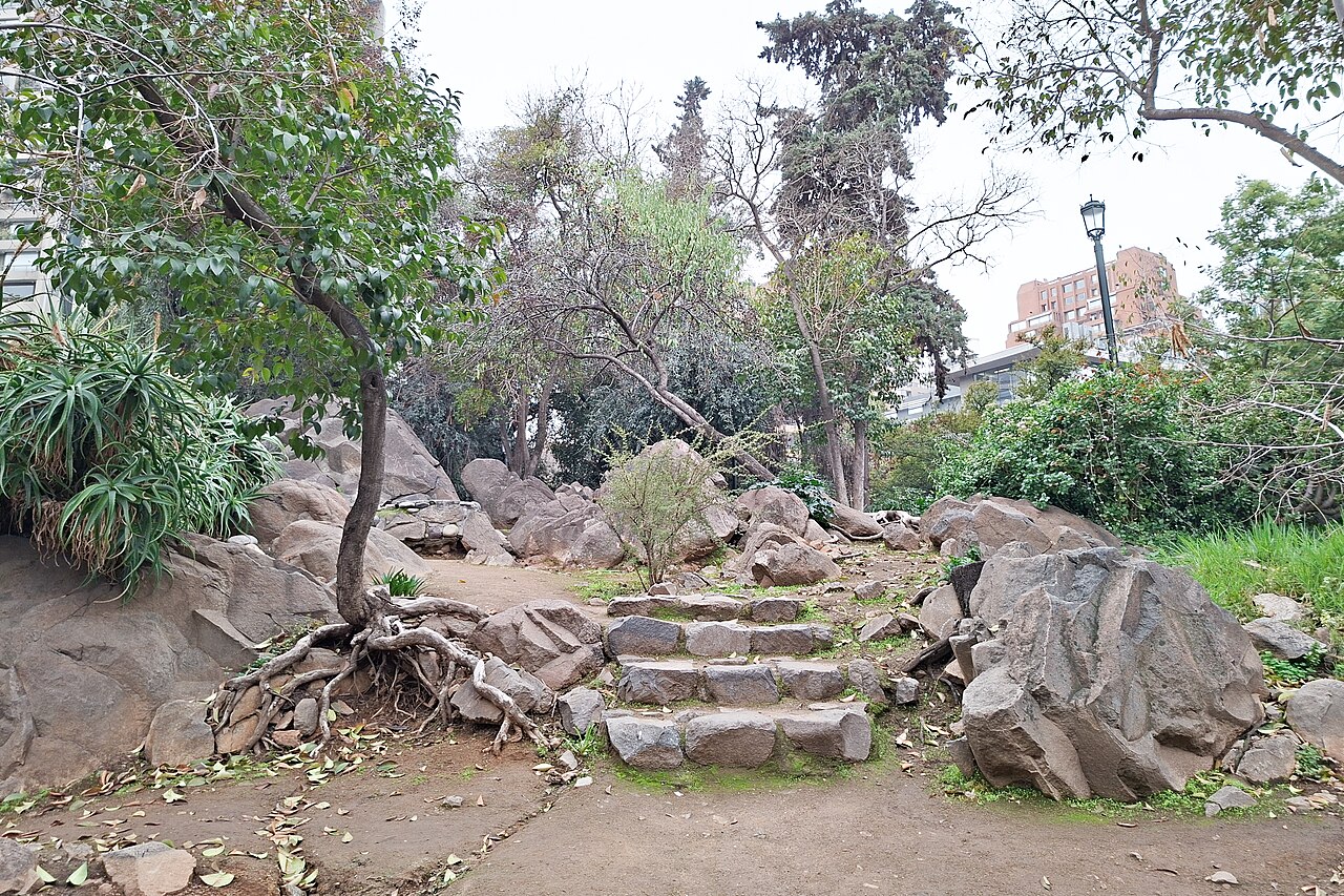 Stone steps and large boulders on a tree-lined trail on Cerro Navidad, a small hill in Las Condes, with apartment buildings behind
