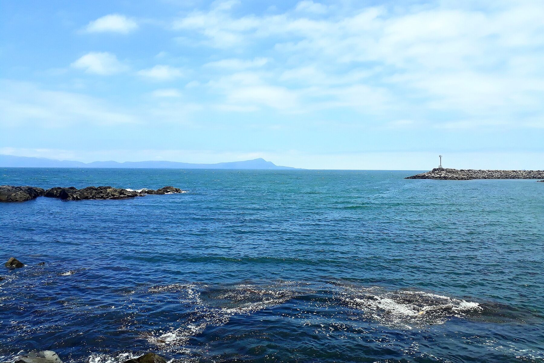 Blue Pacific water and dark rocks at Playa San Miguel, with a stone jetty and a distant headland under a light blue sky