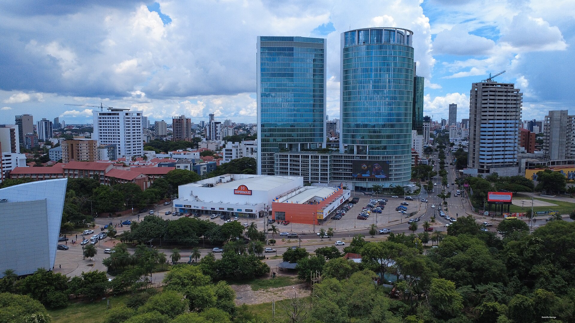 Aerial view of two glass office towers behind a white supermarket with a car park, surrounded by trees and city buildings