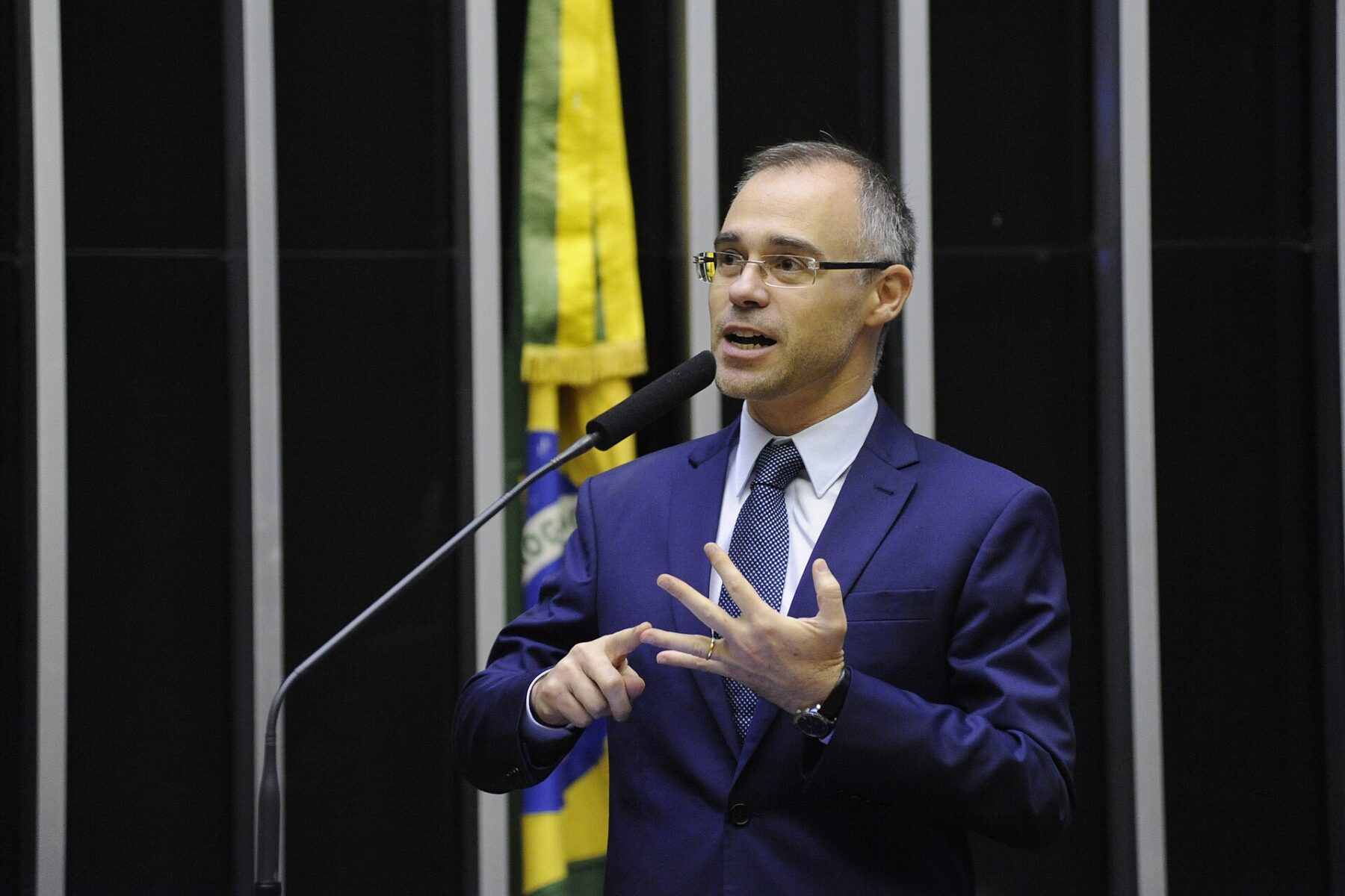 André Mendonça in a blue suit and glasses speaks into a microphone and gestures, with a Brazilian flag behind him