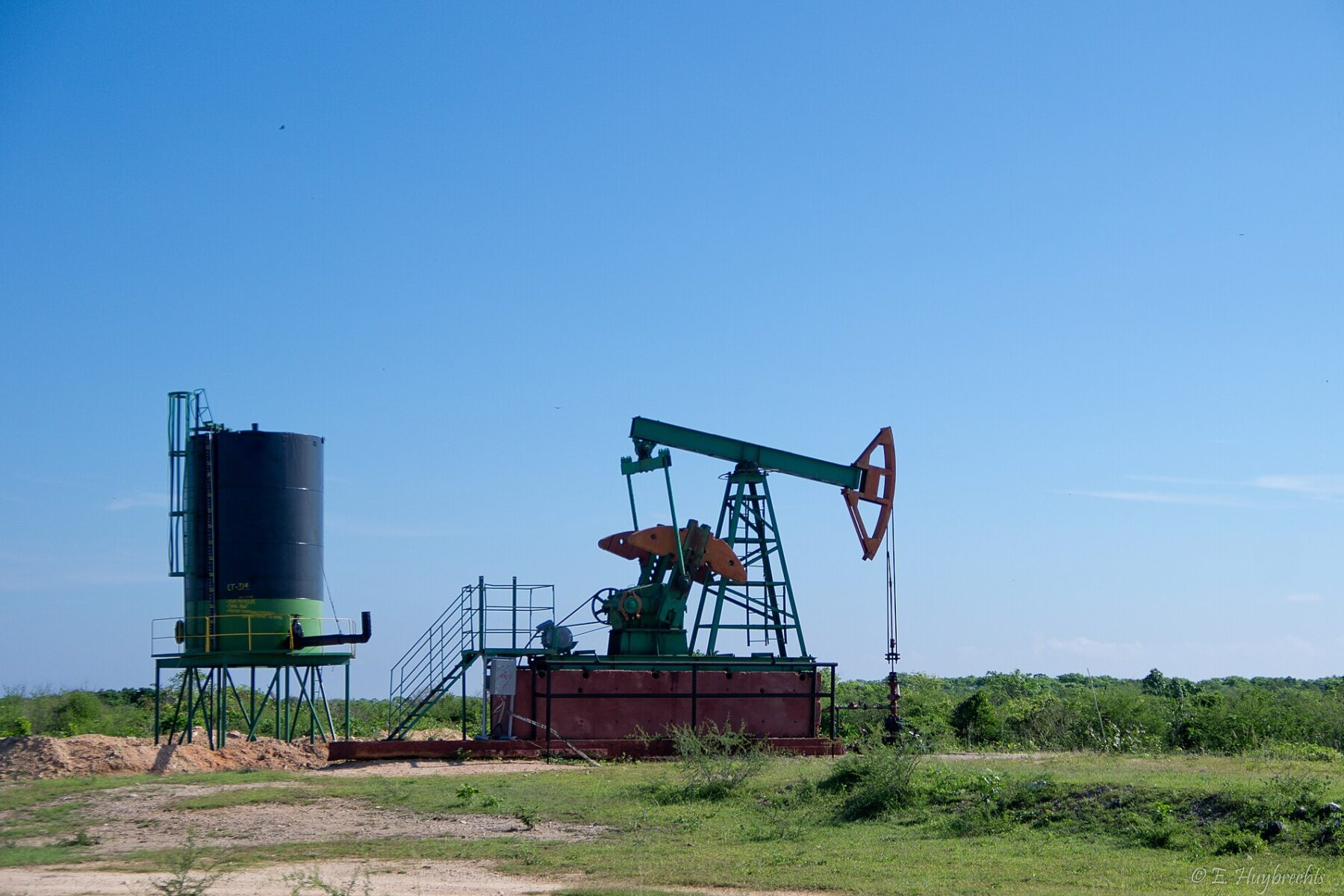 A green and orange oil pumpjack beside a black storage tank on scrubland in Cuba under a clear blue sky