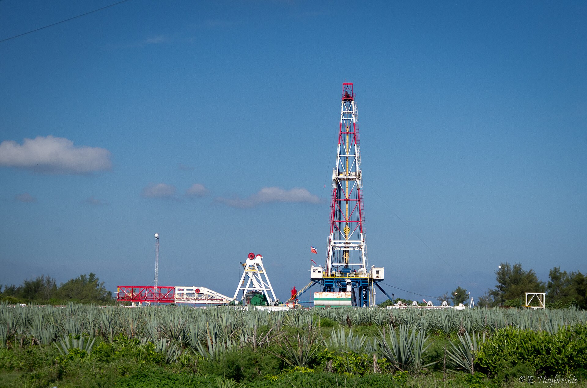 A red and white oil drilling rig with support equipment rising above a field of agave plants in Cuba under a blue sky
