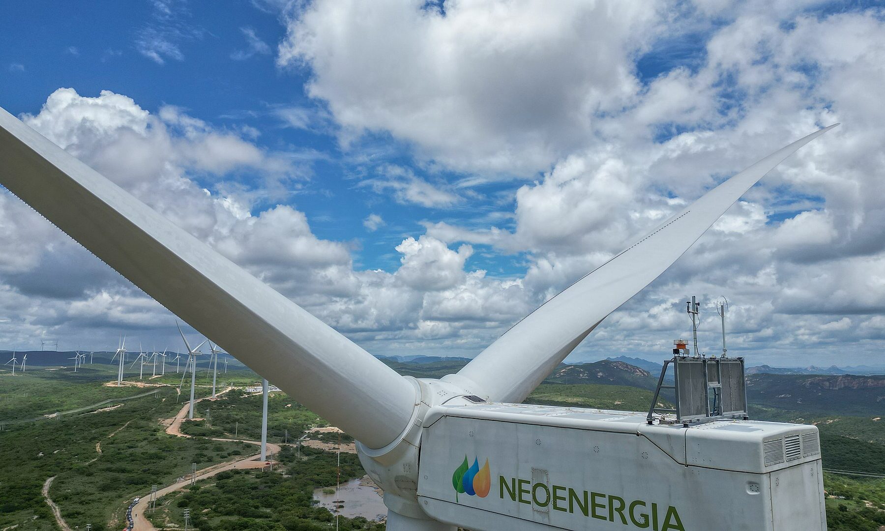 A white Neoenergia wind turbine nacelle and blades above green hills dotted with more turbines under a cloudy blue sky