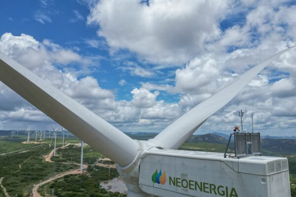A white Neoenergia wind turbine nacelle and blades above green hills dotted with more turbines under a cloudy blue sky