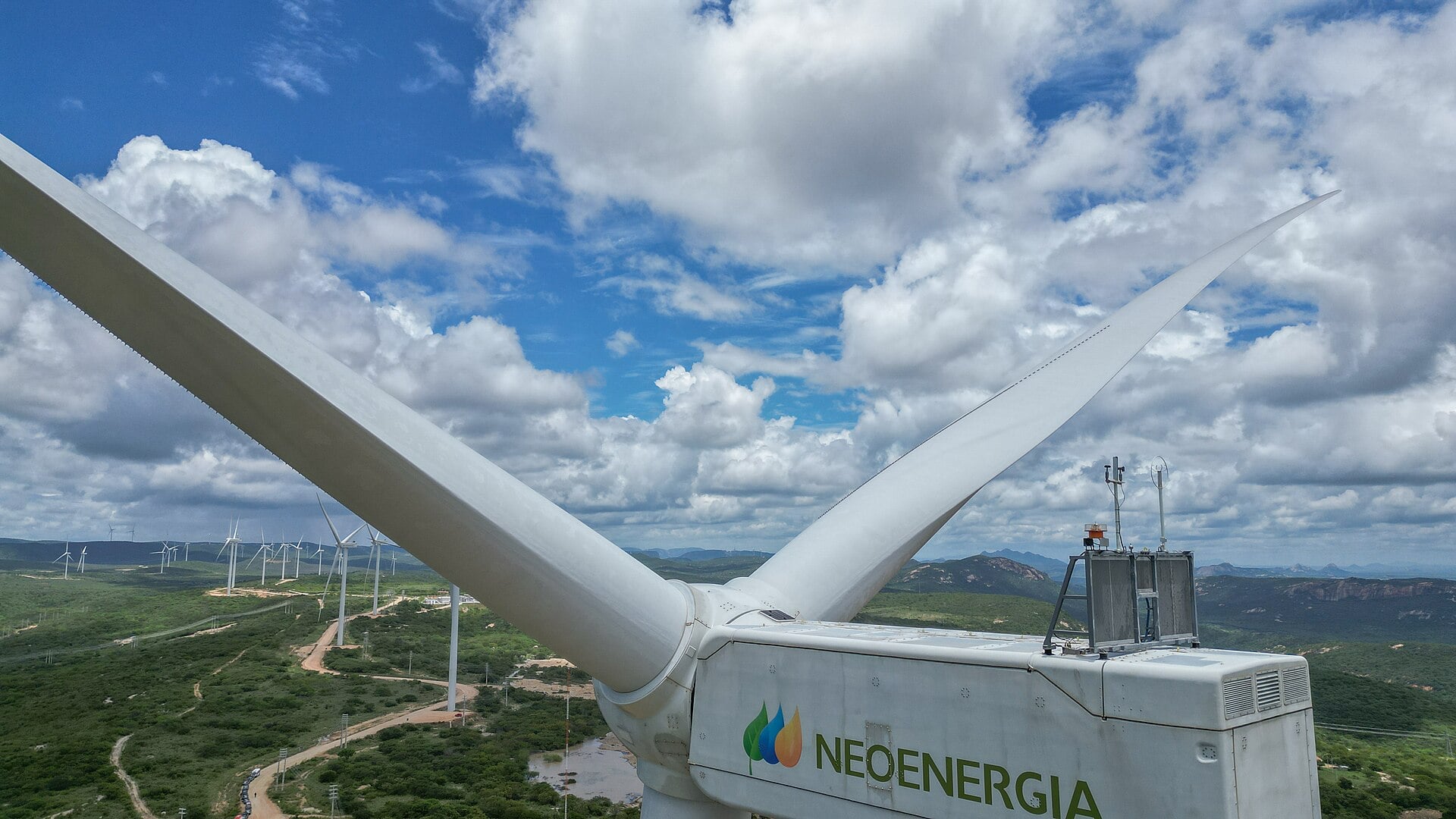 A white Neoenergia wind turbine nacelle and blades above green hills dotted with more turbines under a cloudy blue sky