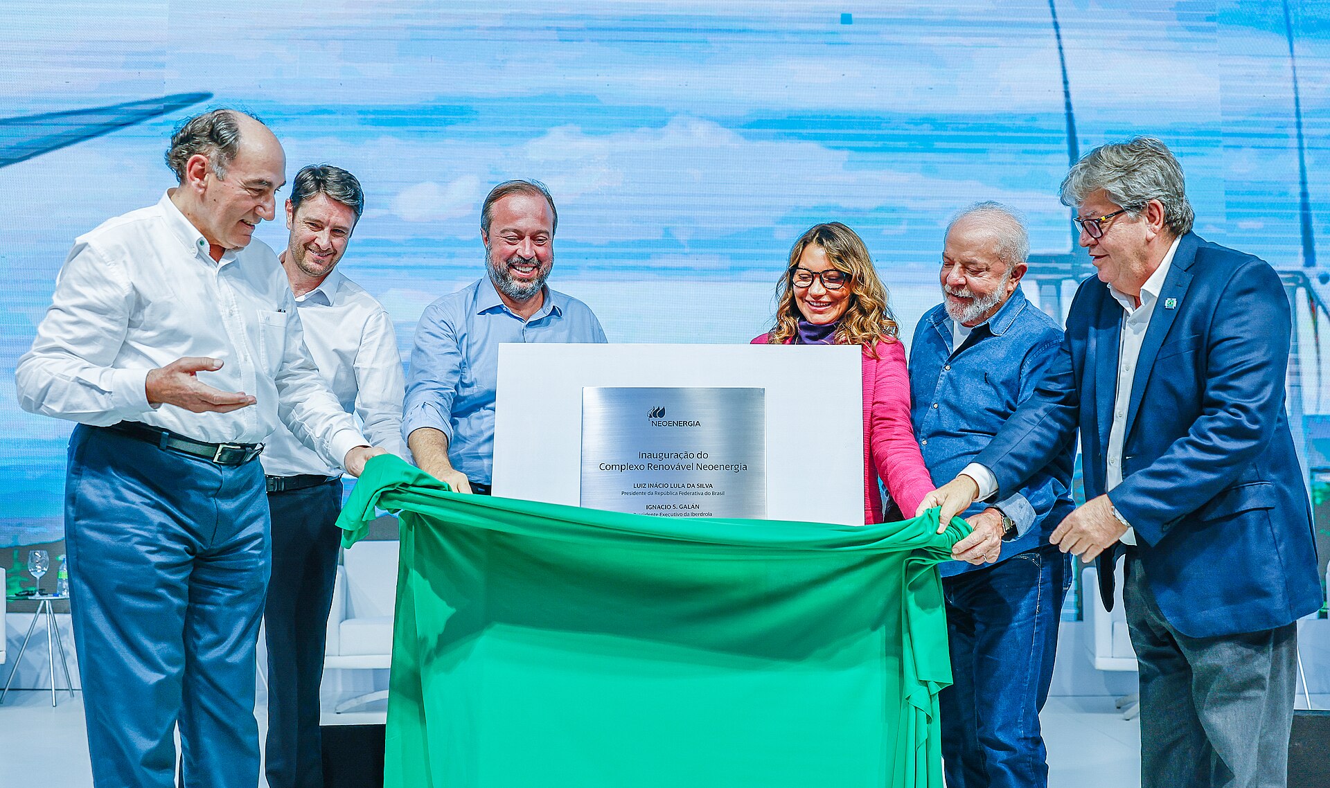 Six officials pull back a green cloth to unveil a Neoenergia plaque at a ceremony, with wind turbines on the backdrop behind them