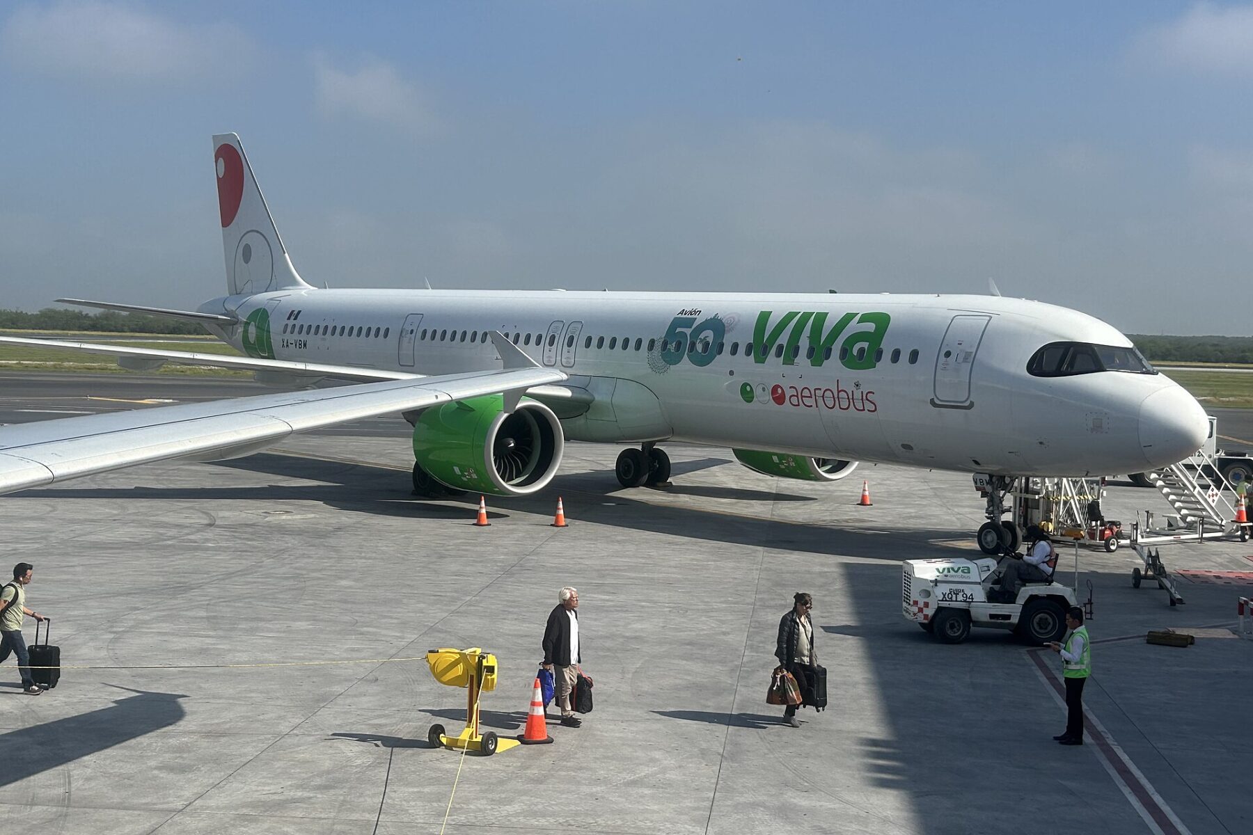 A white Viva Aerobus Airbus A321neo parked at a Monterrey airport stand with passengers carrying bags and a ground crew worker on the apron