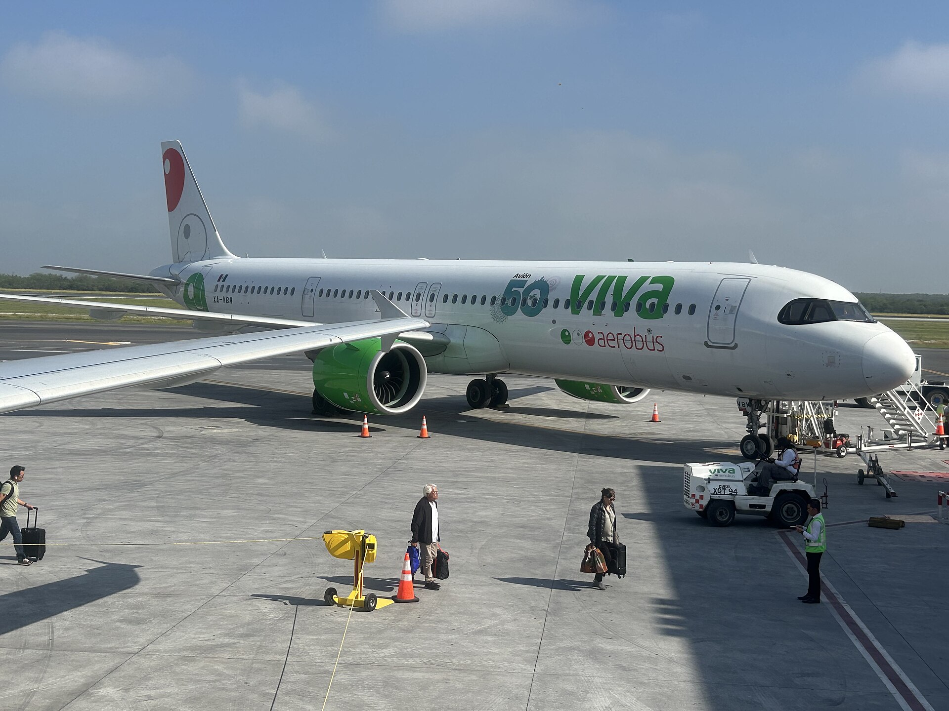 A white Viva Aerobus Airbus A321neo parked at a Monterrey airport stand with passengers carrying bags and a ground crew worker on the apron