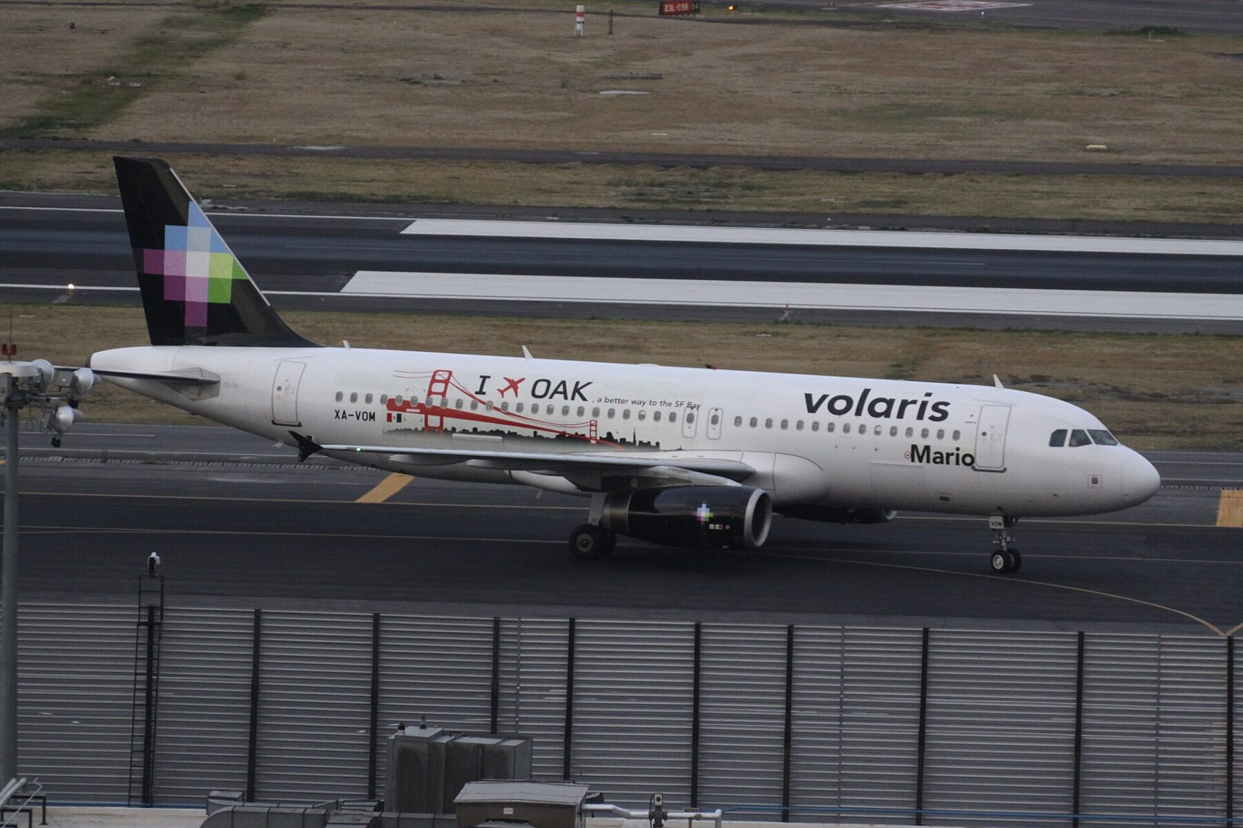A white Volaris Airbus A320 with a black tail and a pixel-pattern logo taxiing on a runway at Mexico City International Airport