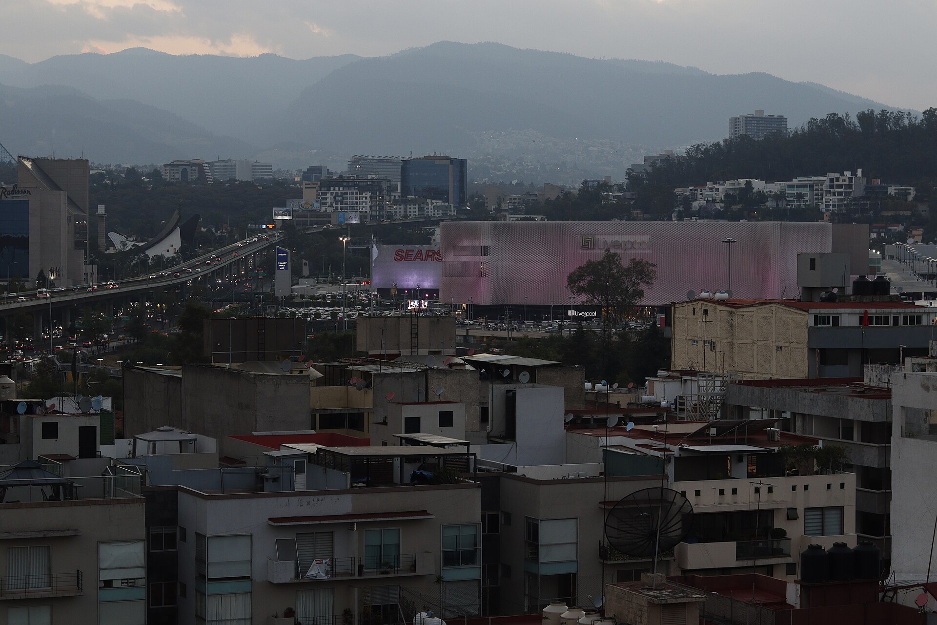 The Perisur shopping centre at dusk in southern Mexico City, a large illuminated mall building among apartment blocks with mountains behind