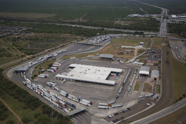 Aerial view of a border crossing with long lines of white semi-trailer trucks queued beside a large inspection building and a river in the background
