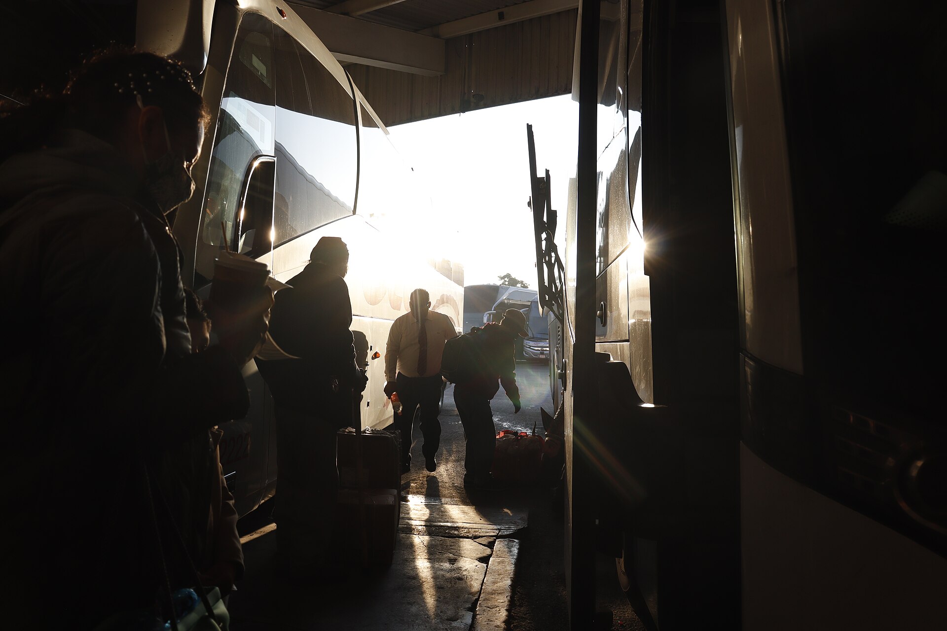 Silhouetted passengers carry bags between two coaches in a bus terminal bay, with low sun glaring through the open end