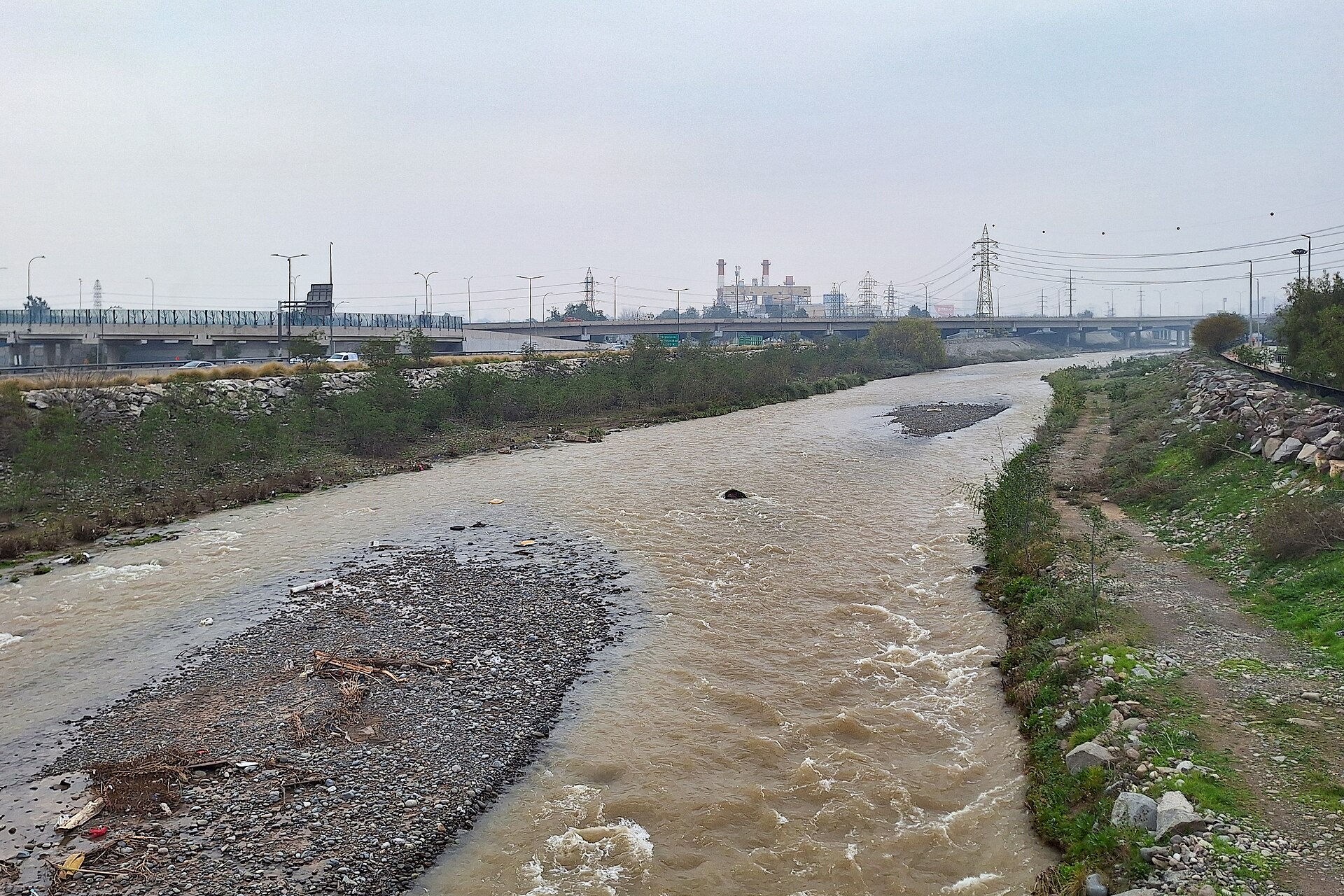 Brown, fast flowing Mapocho River between gravel banks and highway bridges under an overcast sky in Santiago