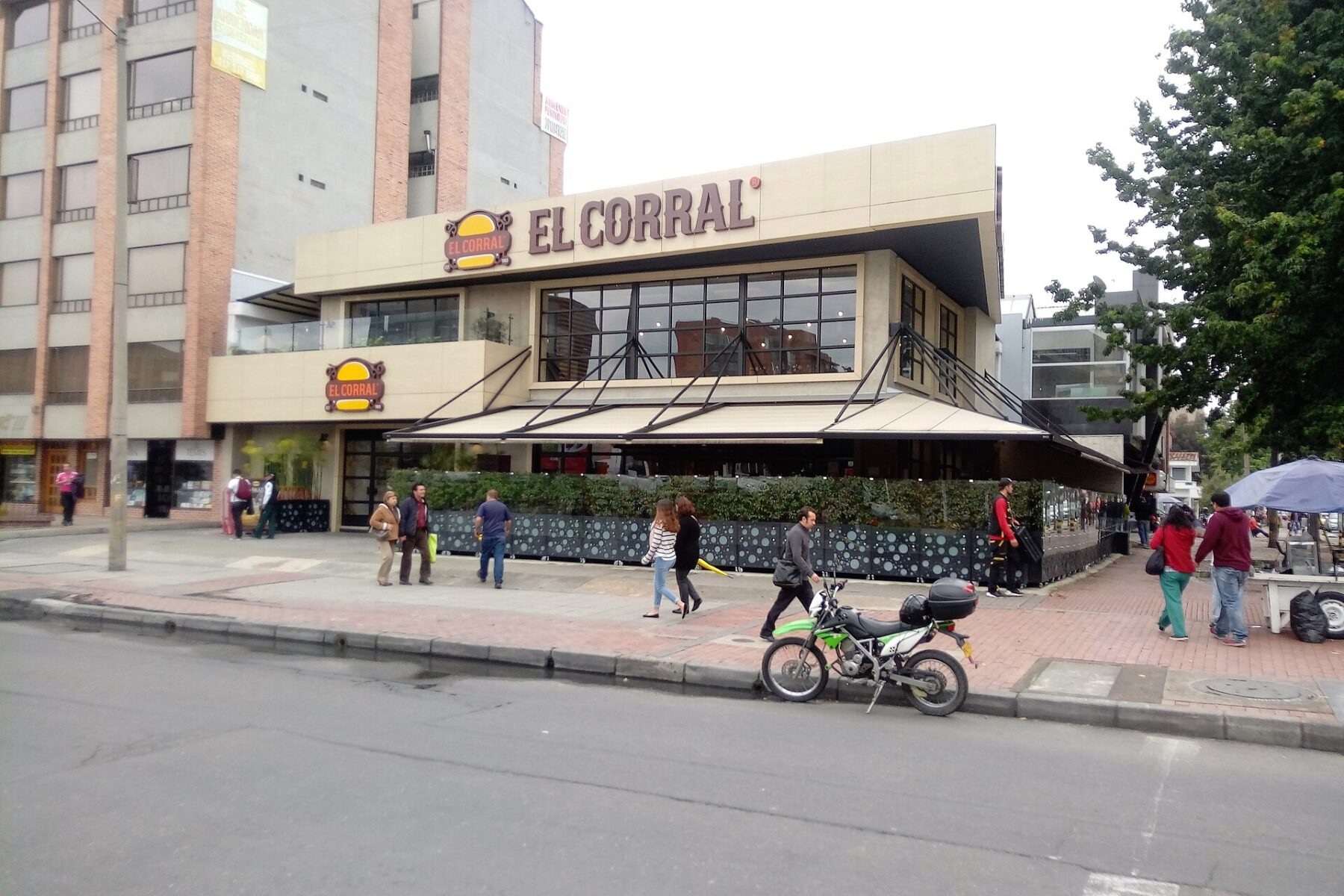 Two-storey El Corral burger restaurant with a brown sign on a Bogota street corner, pedestrians and a green motorbike on the pavement under an overcast sky