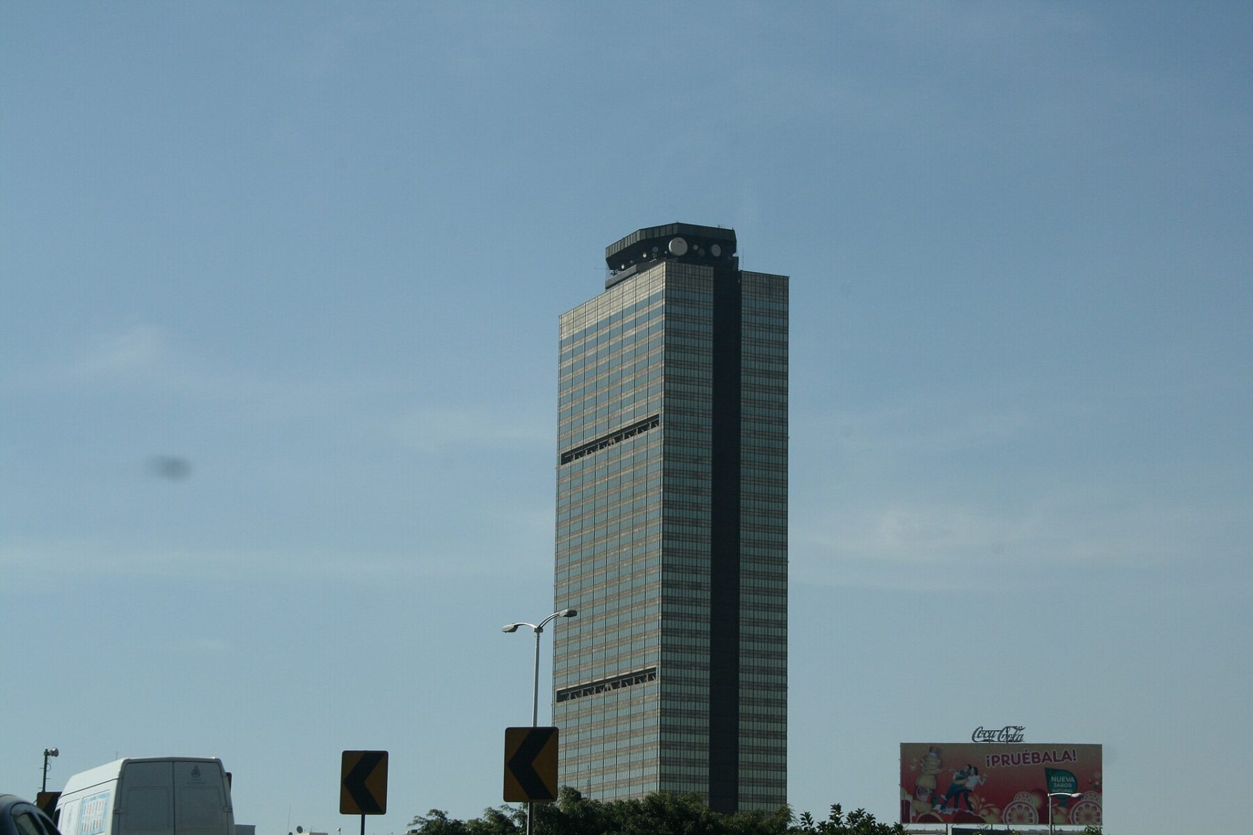 The Pemex headquarters tower in Mexico City rising above traffic signs and a van under a hazy blue sky