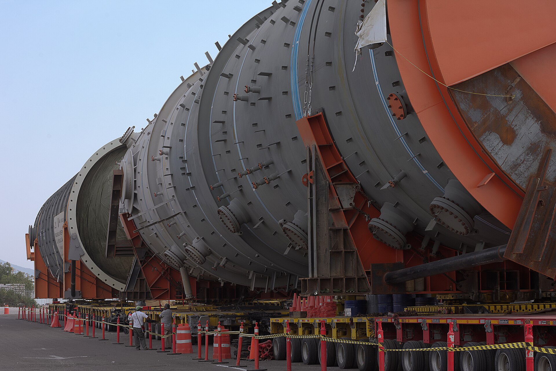 Large grey and orange distillation tower sections lying on heavy transport trailers beside orange traffic cones