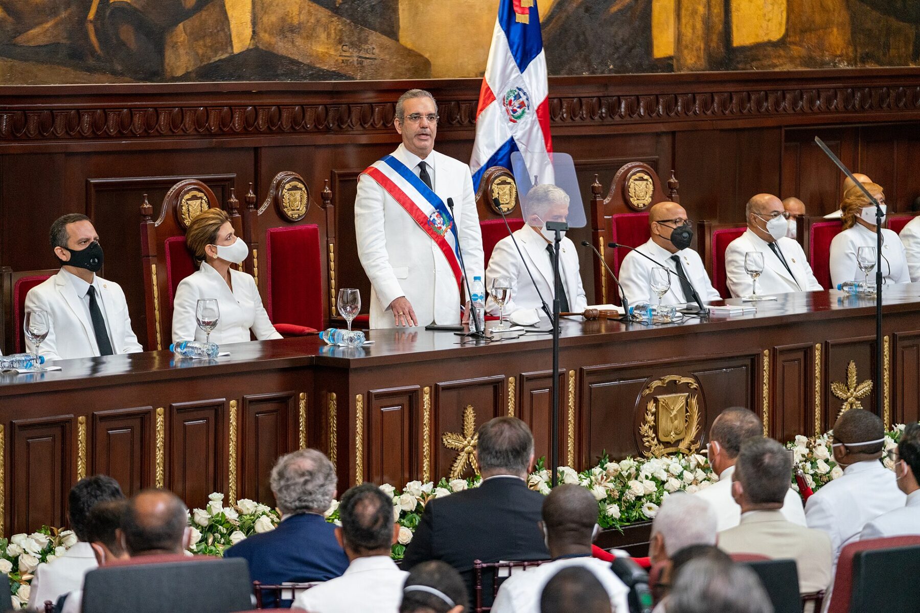 Luis Abinader in a white suit and presidential sash speaks at a lectern in Congress, with officials seated behind him