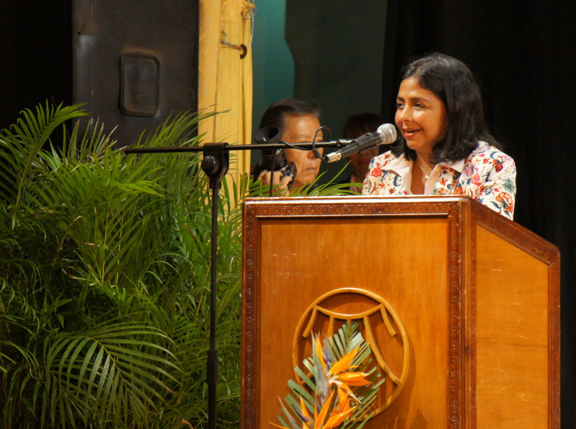 Delcy Rodríguez in a floral jacket smiles while speaking into a microphone at a wooden lectern beside palm leaves