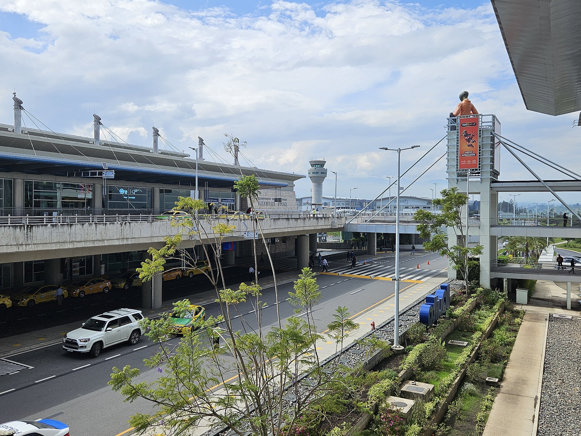 The glass terminal building and control tower of Quito airport seen from an upper walkway, with taxis on the access road under a cloudy sky