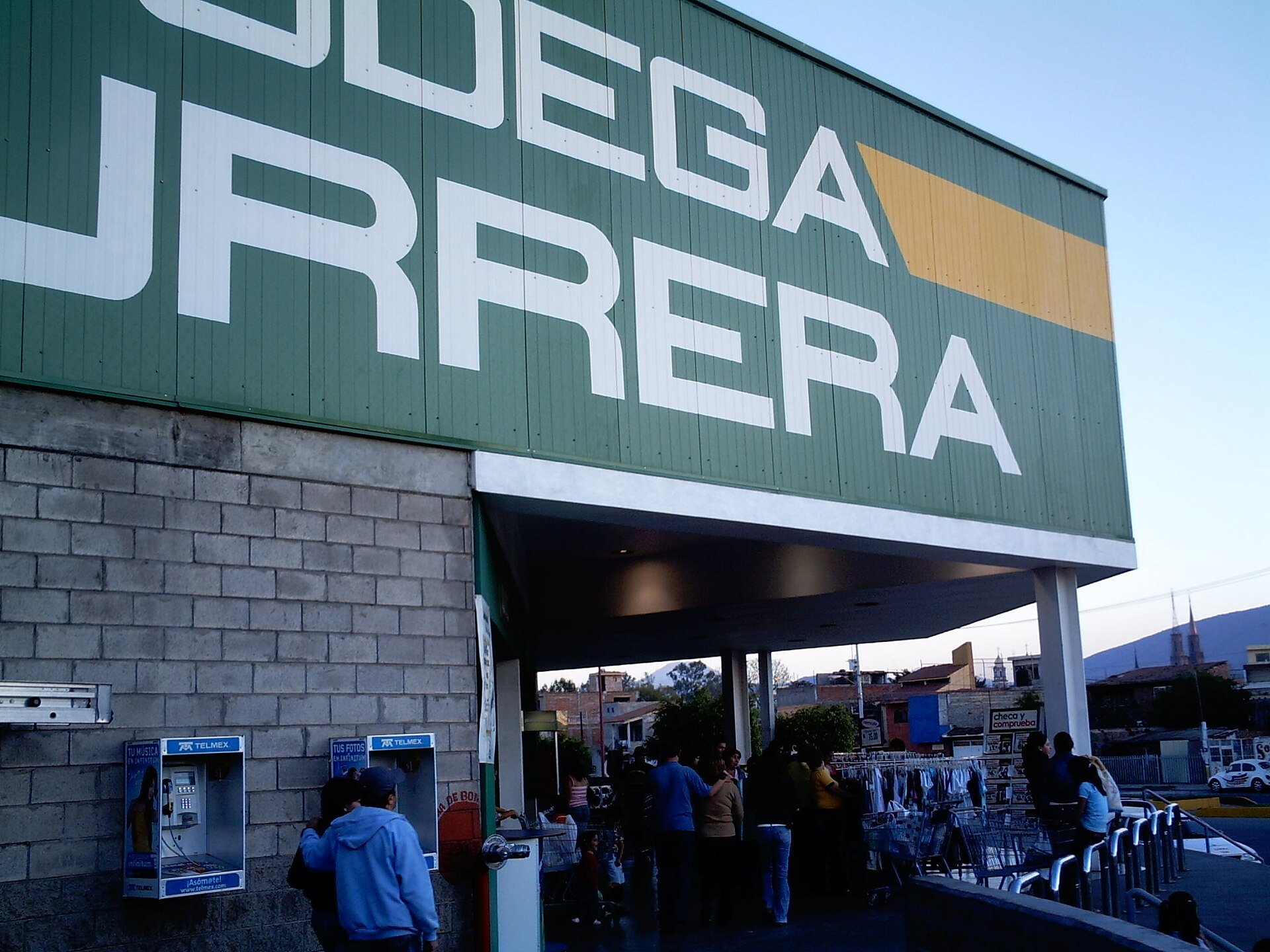 The green Bodega Aurrera sign above a store entrance with shoppers and a pair of payphones, at dusk