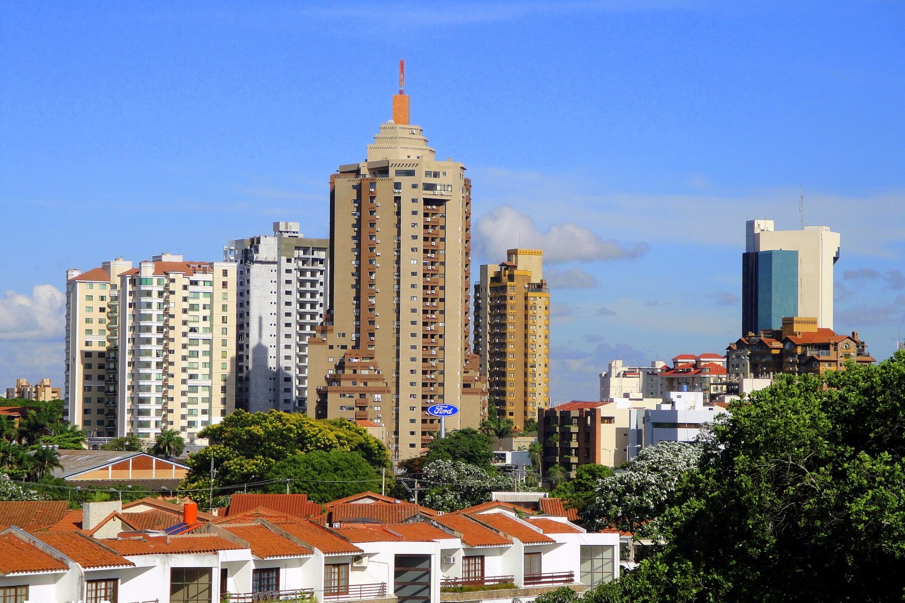 High-rise residential towers and a glass office tower above red-tiled roofs and trees under a blue sky