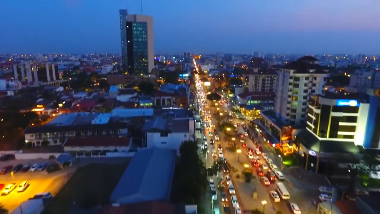 Aerial dusk view of a city avenue full of car lights, with a tall glass tower on the left and lit buildings on the right