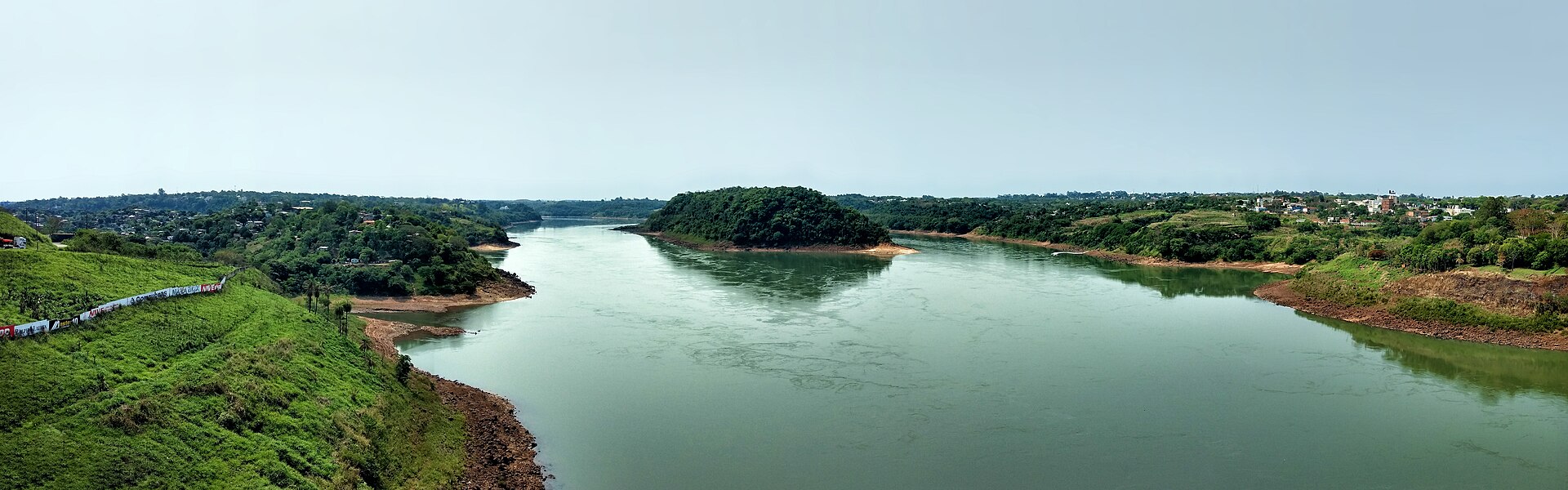 Panoramic view of a wide green river winding between wooded banks and a small forested island, with houses on a hill to the right