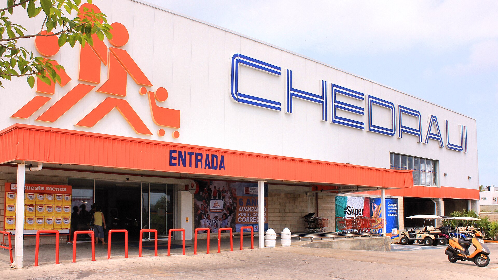 The entrance of a Chedraui supermarket in Isla Mujeres, Mexico, with the orange shopper logo, blue Chedraui lettering and a motorbike and golf carts outside