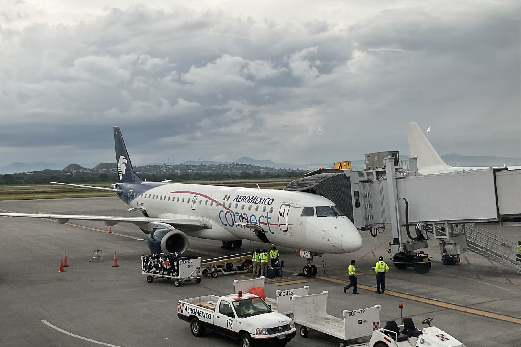 An Aeromexico Connect Embraer 190 jet at a jet bridge with ground crew and baggage carts under a cloudy sky
