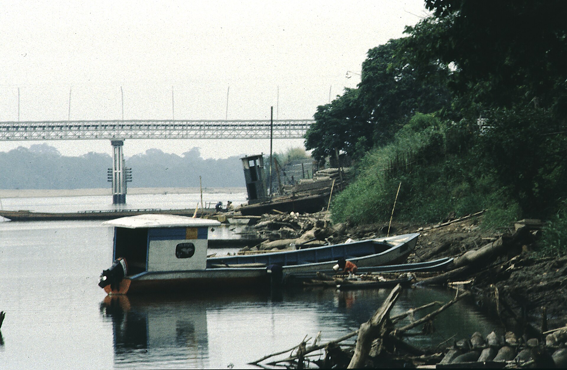 Small river boats moored at a muddy bank of the Napo River at Coca, with a long truss bridge and hazy hills behind