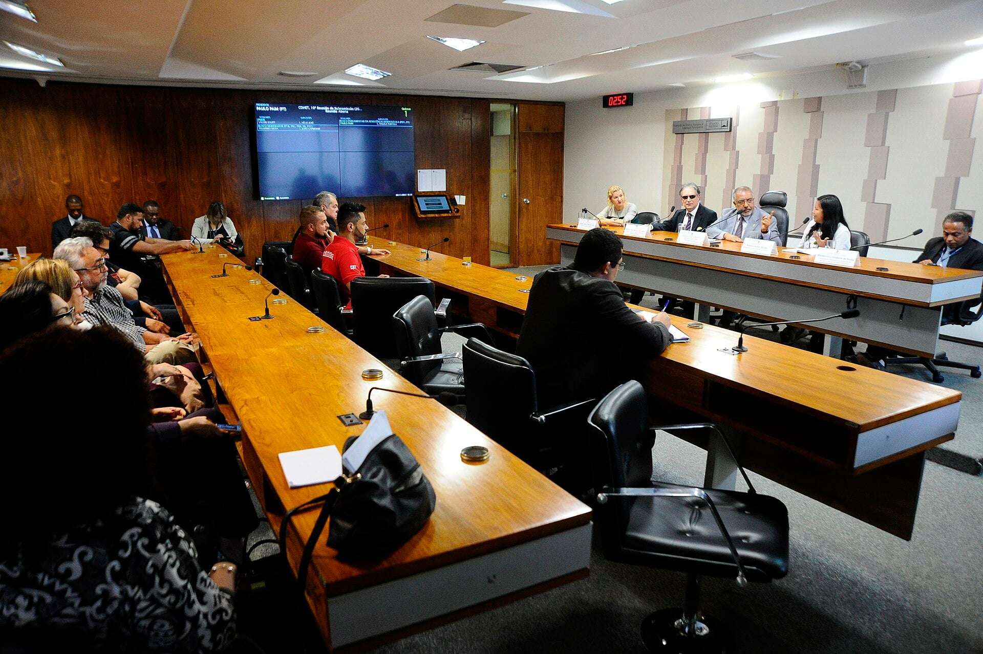 A committee room in Brasília with delegates seated along wooden tables and a screen on the wall during a Senate subcommittee hearing