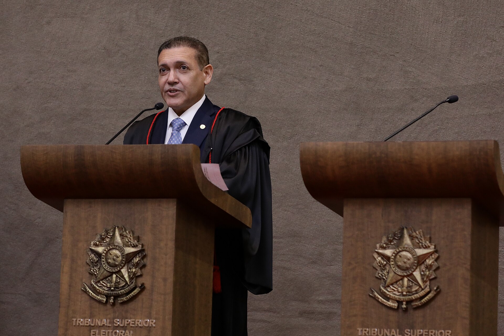 Justice Kassio Nunes Marques in judicial robes speaking at a wooden lectern marked Tribunal Superior Eleitoral, a second lectern beside him