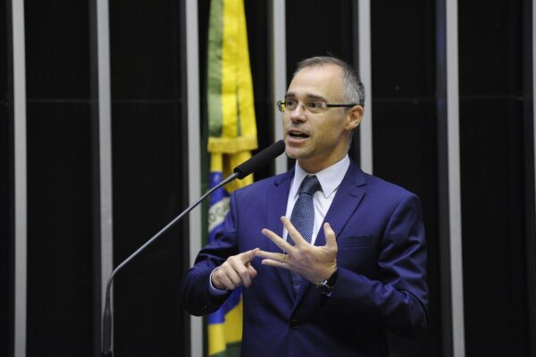 André Mendonça in a blue suit and glasses speaks into a microphone and gestures, with a Brazilian flag behind him
