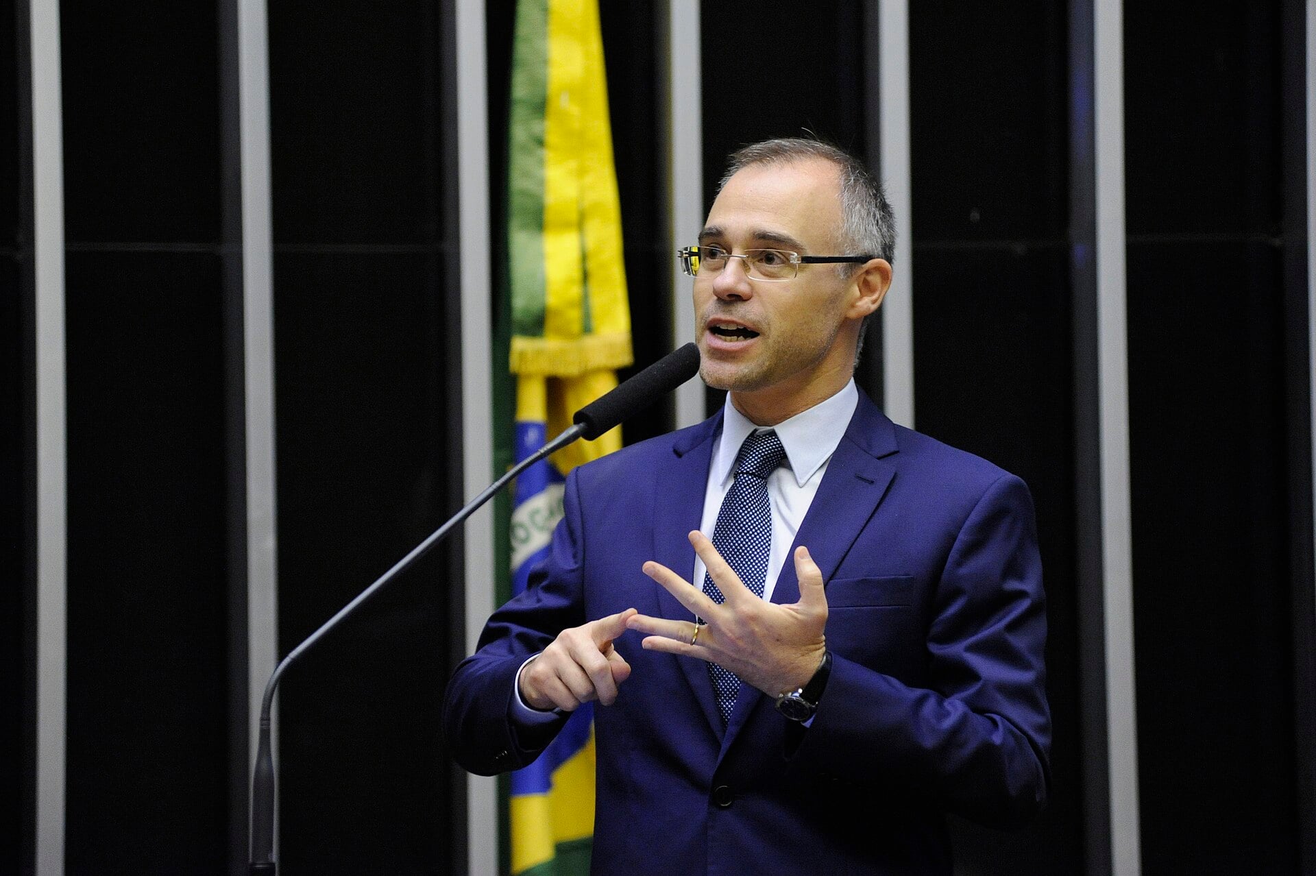 André Mendonça in a blue suit and glasses speaks into a microphone and gestures, with a Brazilian flag behind him