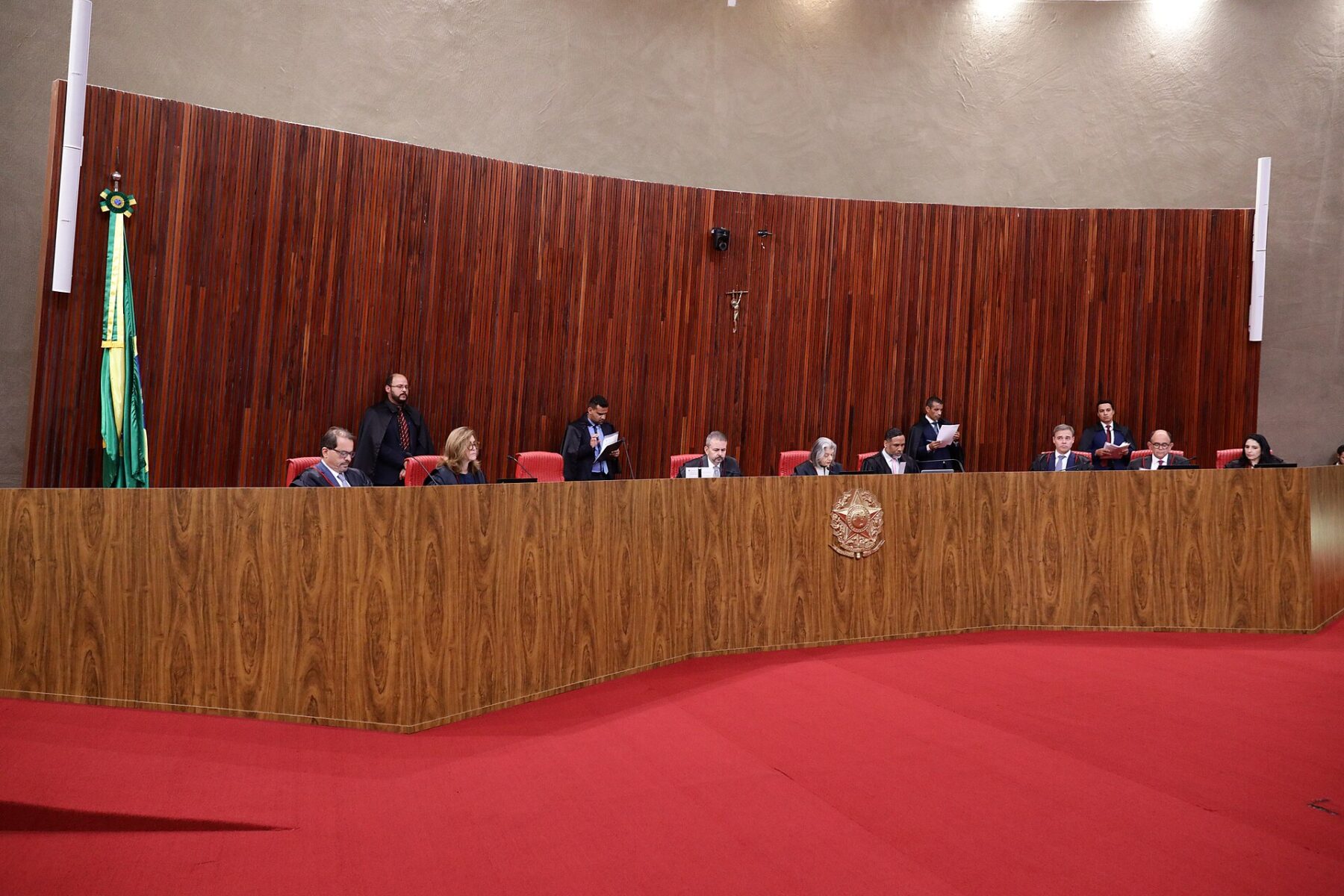 Wide view of the Superior Electoral Court plenary in session, judges seated at a curved wooden bench with the Brazilian flag and a red carpet in front