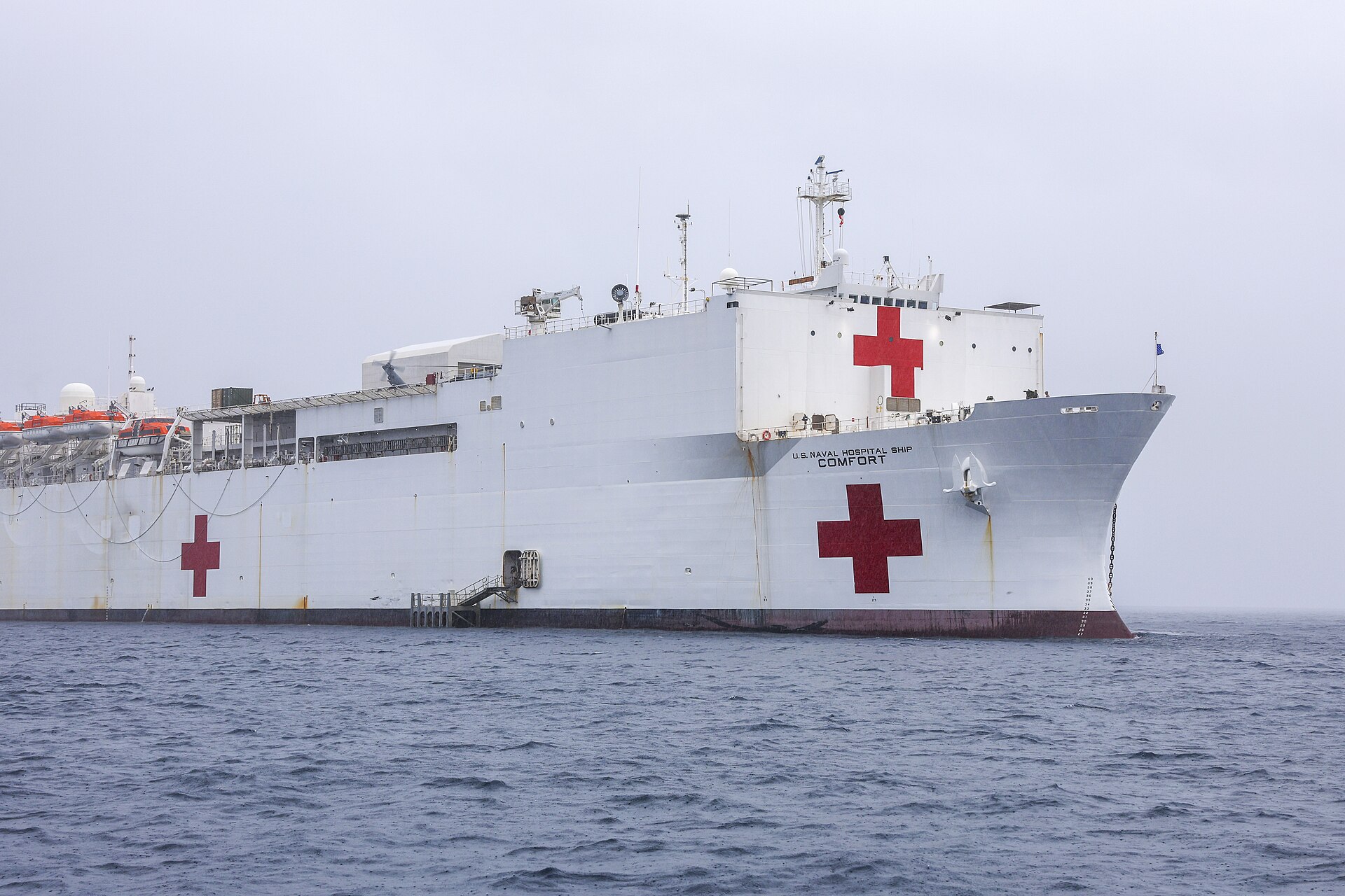 The white US Navy hospital ship USNS Comfort with red crosses on its hull anchored on grey sea under an overcast sky