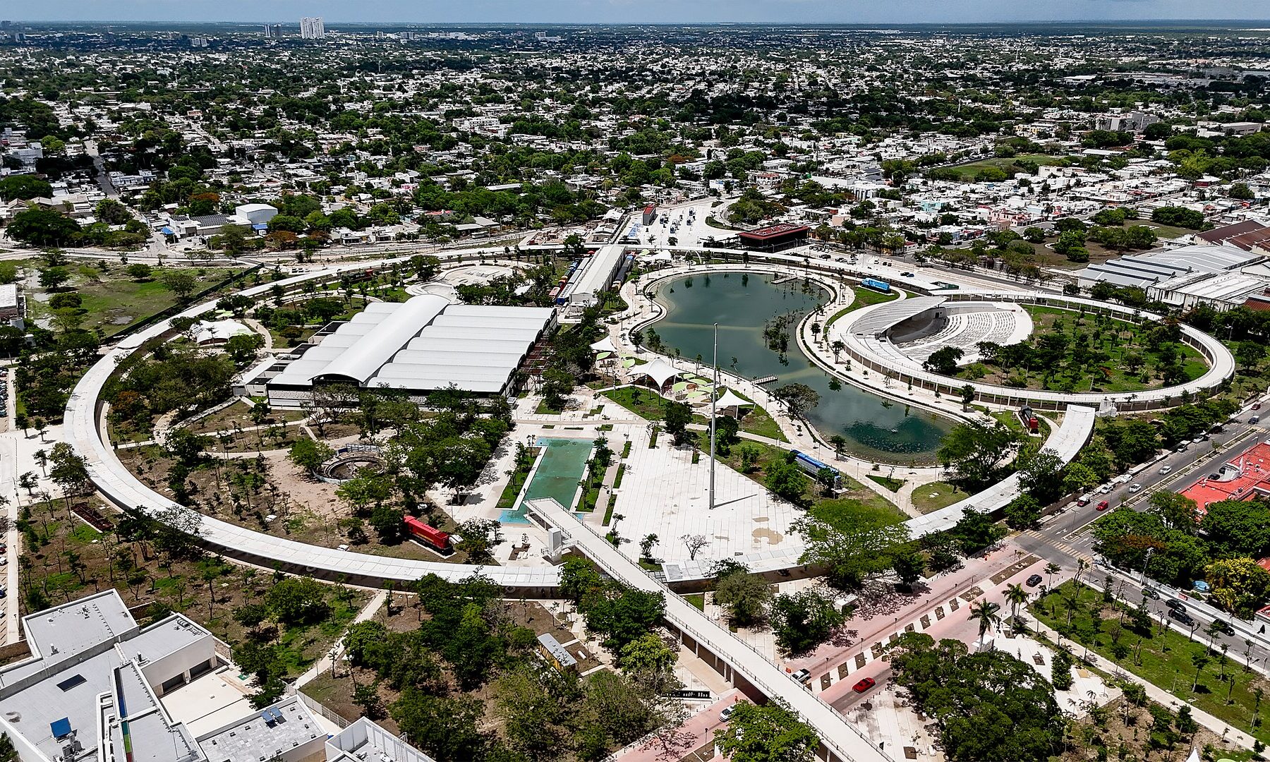Aerial view of a large city park in Mérida with a curved lake, a ring-shaped walkway and an amphitheatre, surrounded by houses and trees