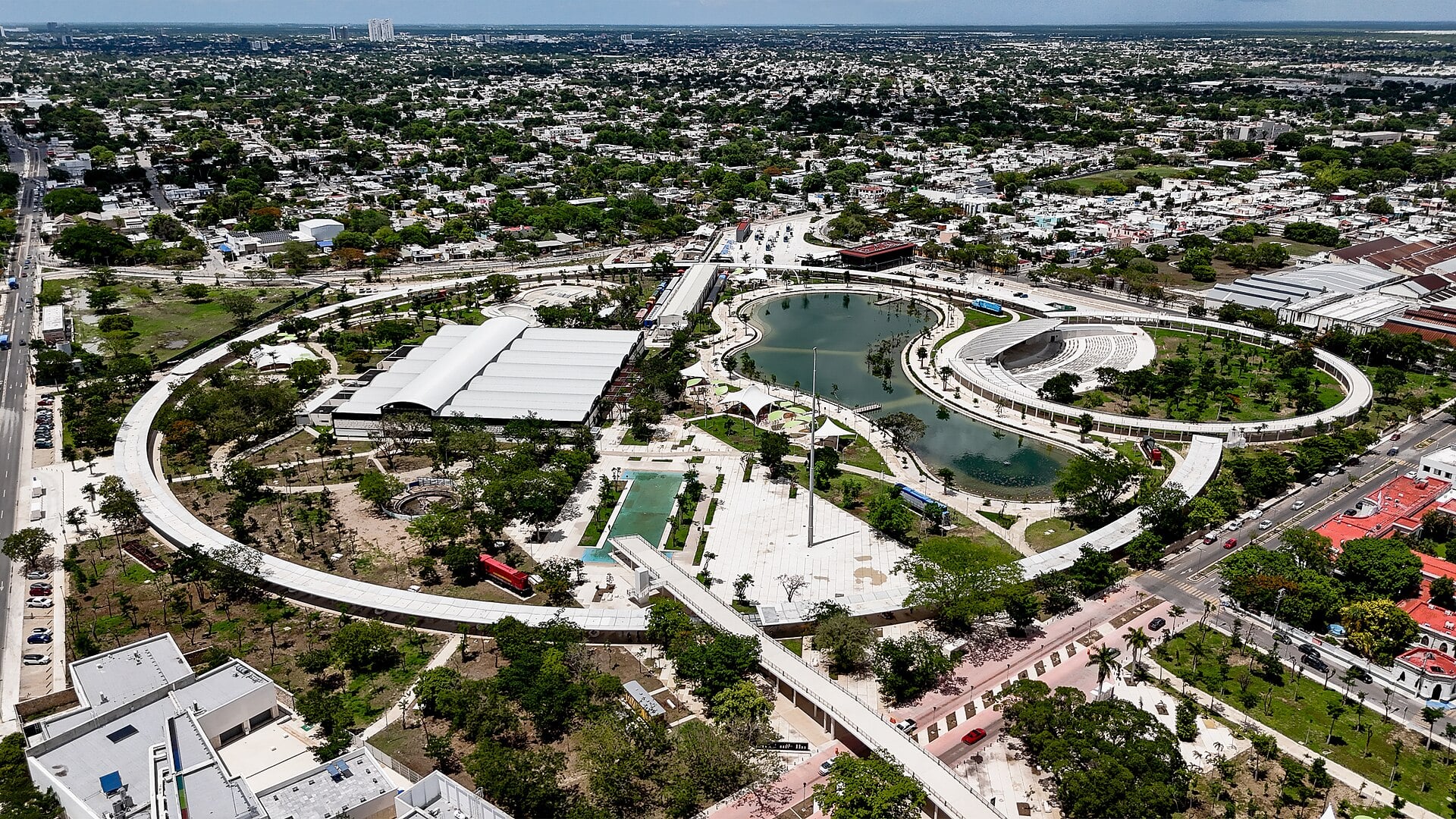 Aerial view of a large city park in Mérida with a curved lake, a ring-shaped walkway and an amphitheatre, surrounded by houses and trees