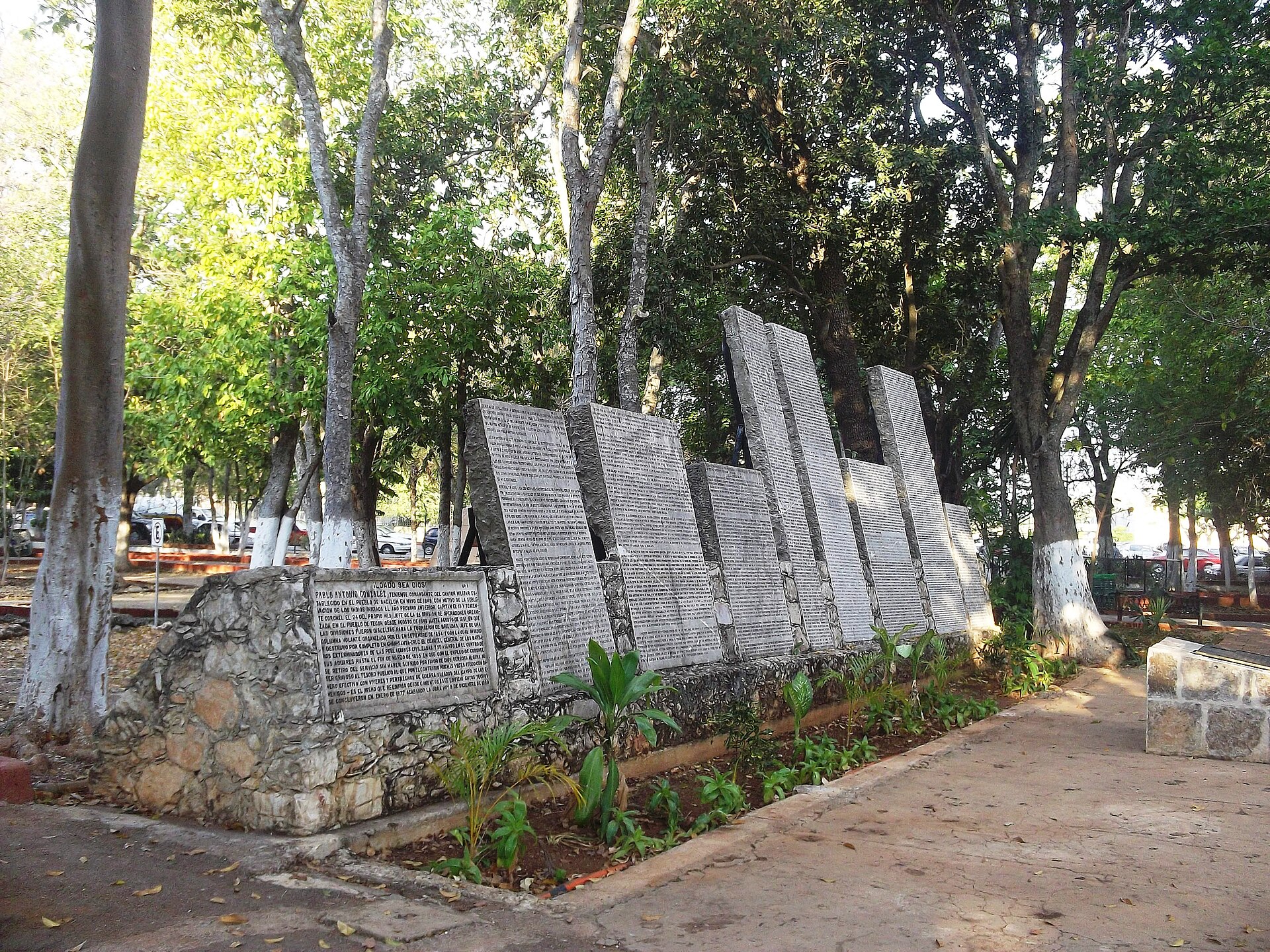 Tall inscribed stone slabs of a monument stand among shaded trees beside a paved path in a Mérida park
