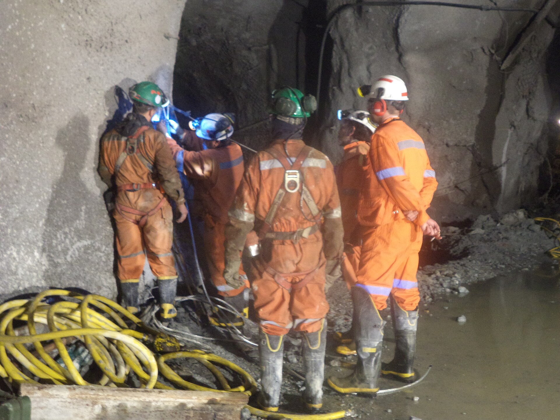 Five miners in orange overalls and helmets work beside a rock wall in a damp tunnel, with yellow hoses coiled on the floor