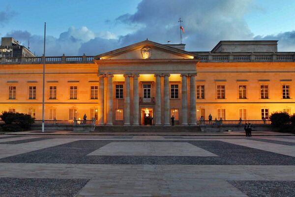 The lit neoclassical facade of the Casa de Nariño presidential palace in Bogotá at dusk, with a wide paved square in front