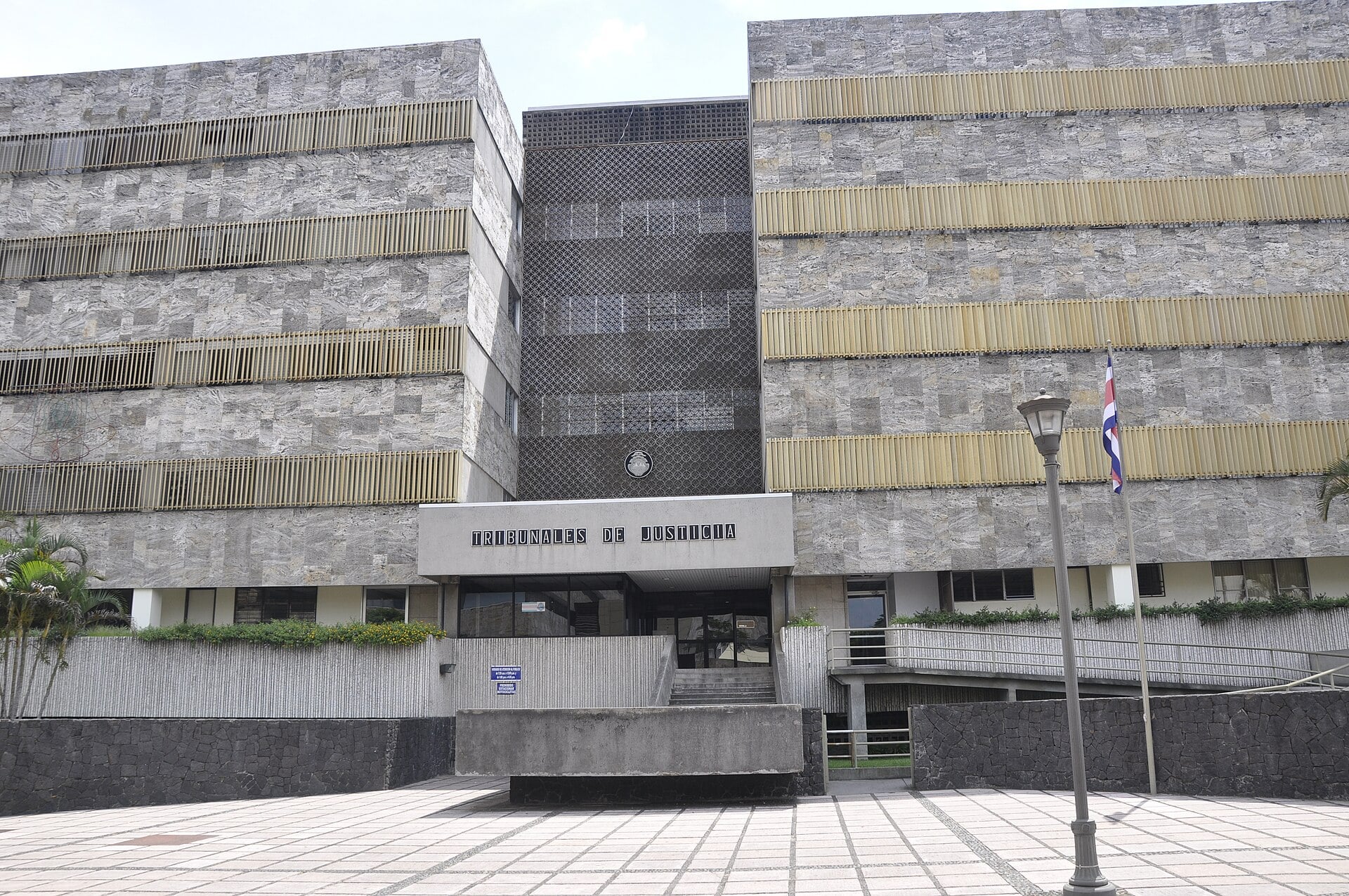 The stone-clad Tribunales de Justicia building in San José with its entrance sign, a Costa Rican flag and a paved plaza in front
