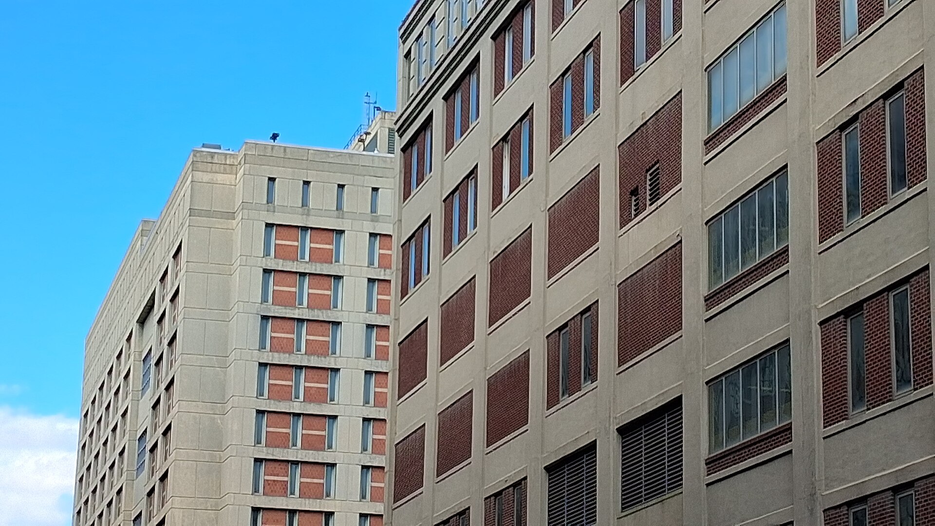 Two tall beige concrete buildings with rows of narrow windows set in red brick panels, rising against a bright blue sky