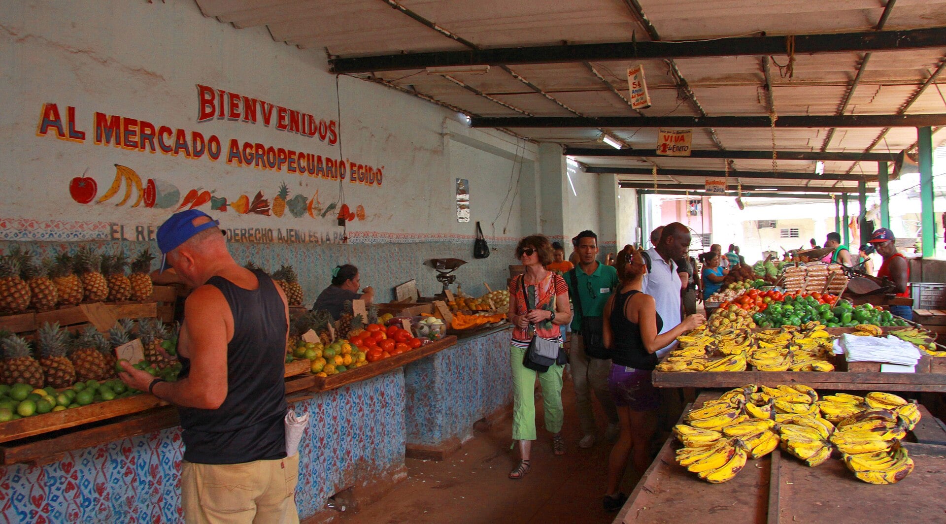 Shoppers and vendors in a covered Havana market hall with piles of pineapples, bananas, tomatoes and peppers and a painted welcome sign