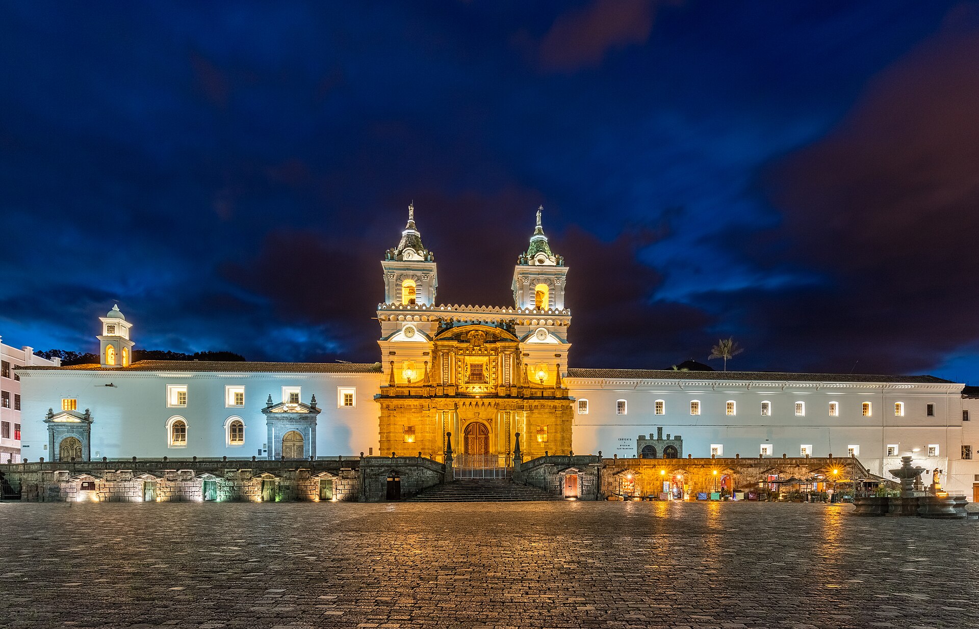 The white facade and twin bell towers of the San Francisco church and monastery in Quito, lit up at dusk above an empty plaza