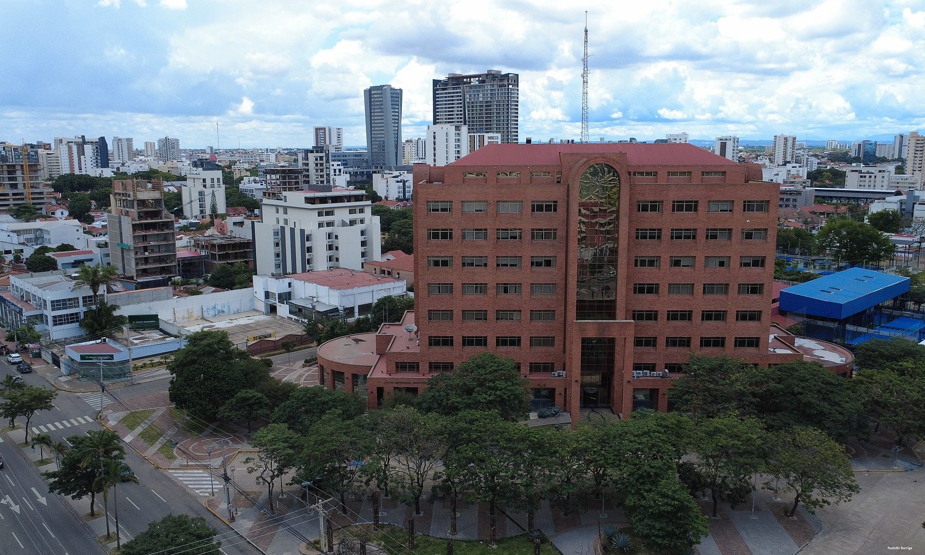 Aerial view of a red-brick office tower surrounded by trees and low buildings, with high-rises and a cloudy sky behind